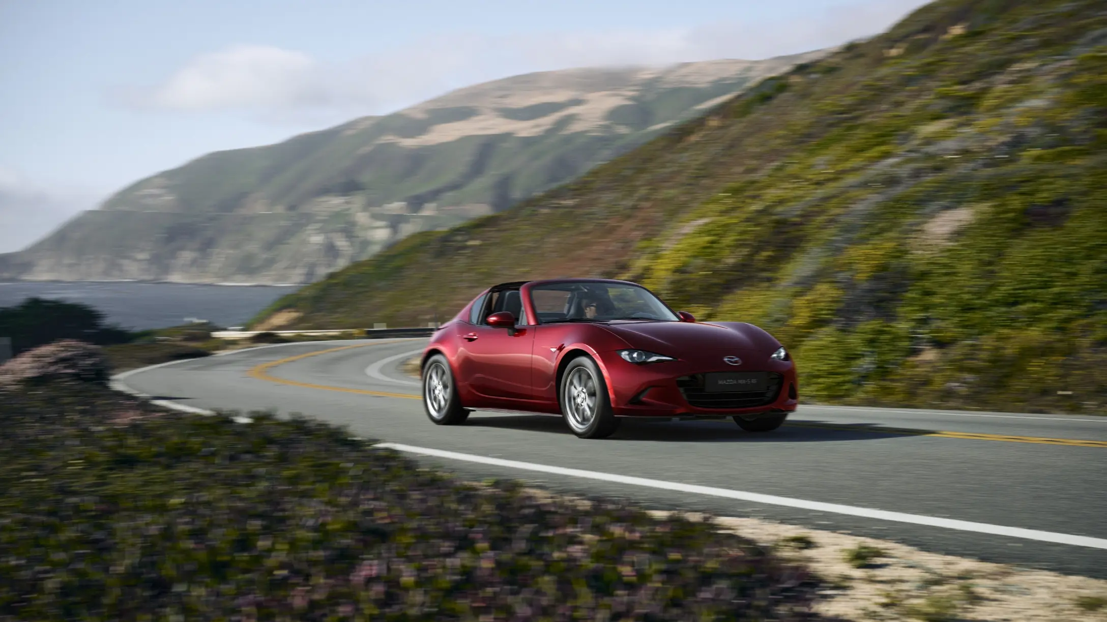 Mazda MX-5 rojo descapotable circula por una carretera costera con montañas de fondo.